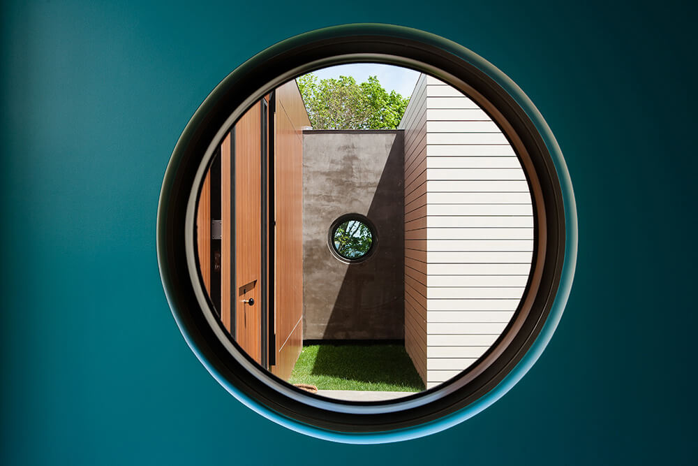CNR Group luxury home construction: View through a large circular window showing a small courtyard with wooden and white siding walls, grass, and a smaller circular window reflecting greenery.