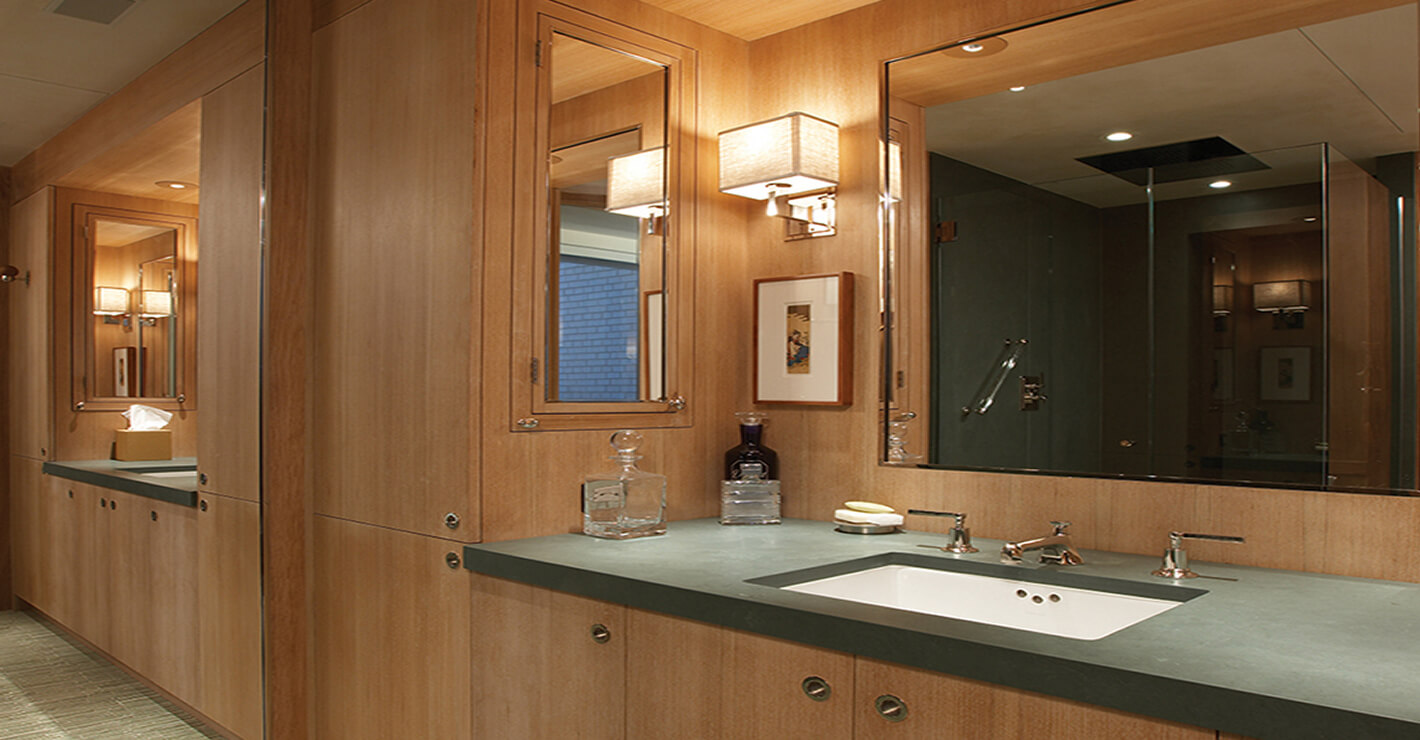 CNR Group luxury home construction interior: Modern bathroom vanity with custom millwork wooden cabinets, green countertop, built-in sink, glass decanters, soap dish, and a large mirror with wall light.