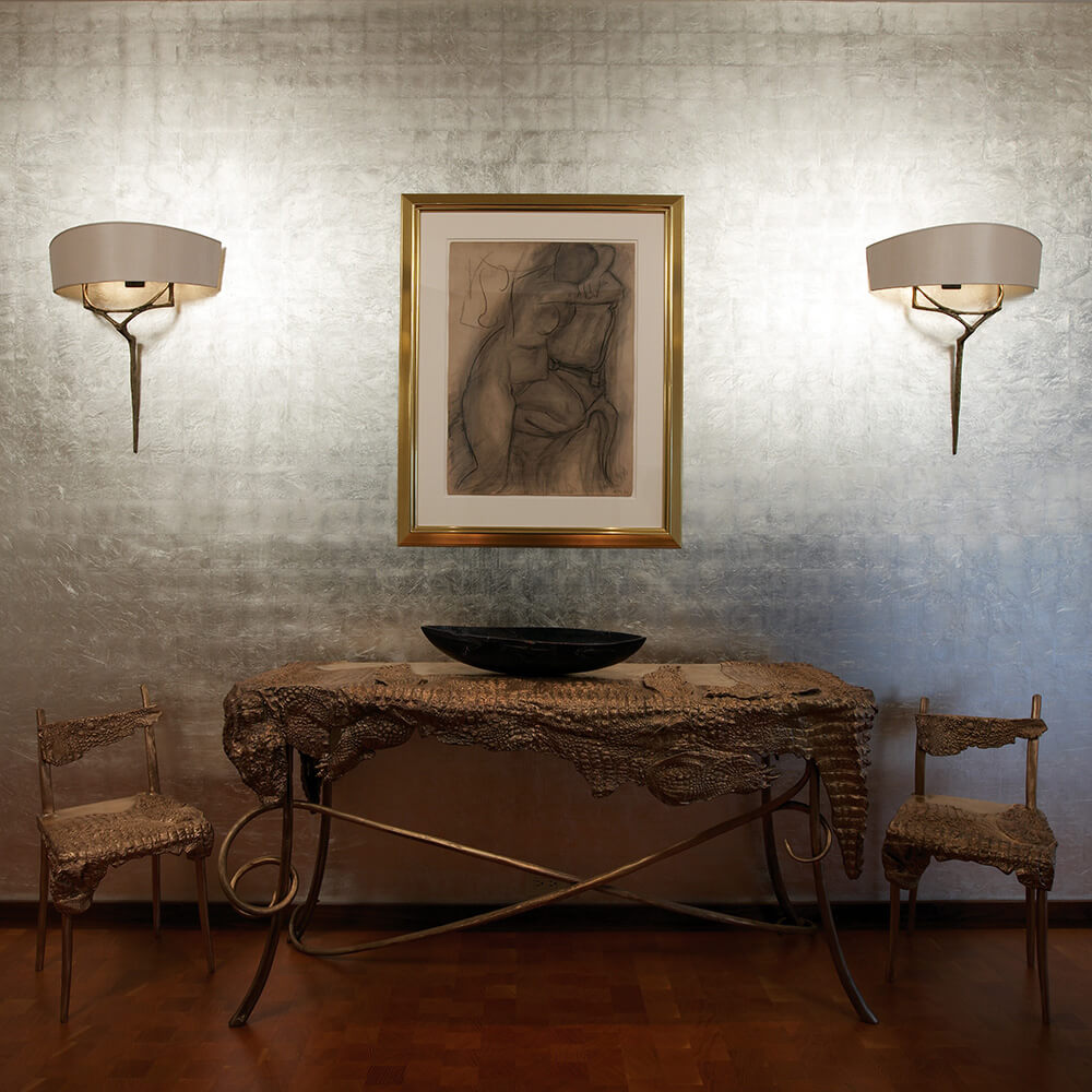 Artistic room with metalwork textured metallic wall, two sculpted chairs, a matching table with a black bowl, and a framed abstract nude drawing between two wall sconces.