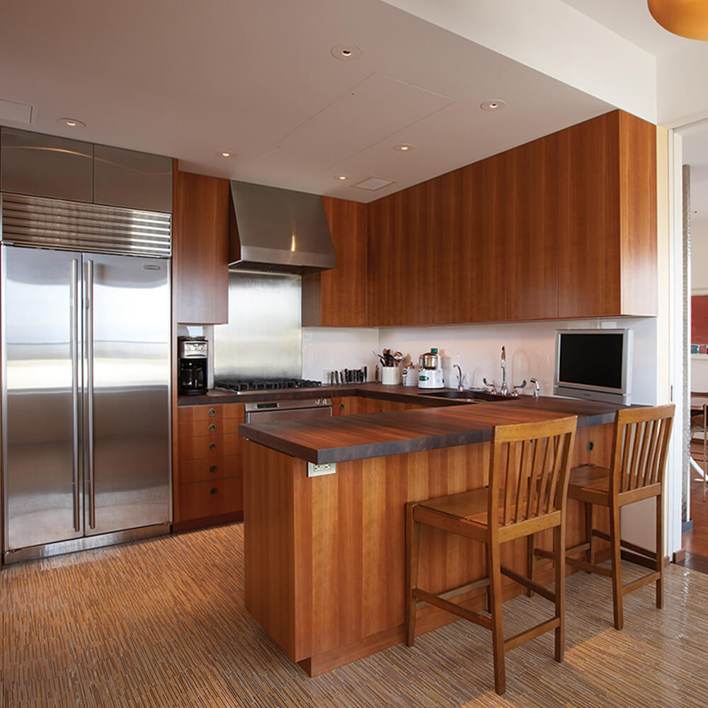 CNR Group NYC luxury home construction interior: Modern kitchen with wood cabinetry, a stainless steel refrigerator, a stovetop with range hood, wood island with two chairs, and a small TV.
