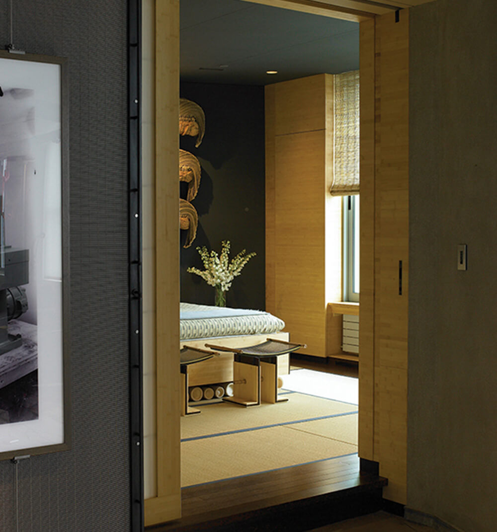 CNR Group luxury home construction interior: View through a sliding wooden door into a minimalist room with tatami mats, a low bed, two stools, and a vase of white flowers.