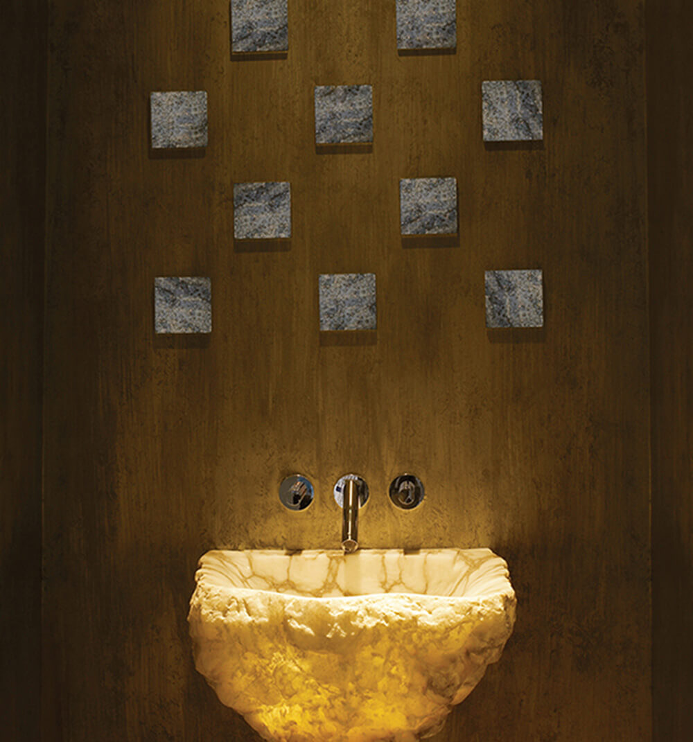 CNR Group luxury home project management interior: Glowing rough stone sink mounted on a textured brown wall with nine square stone tiles above and a modern faucet.
