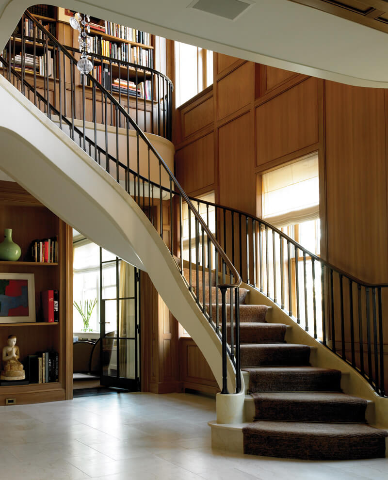 Curved staircase with dark carpeting and black metalwork railing in a wood-paneled room with large windows and built-in bookshelves.