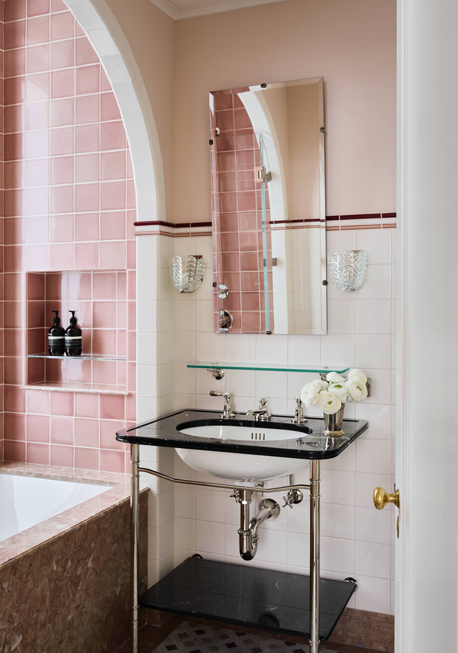 CNR Group NYC luxury home project management interior: Bathroom corner with a black marble sink, white flowers in a silver vase, a rectangular mirror, and pink tiled wall around a bathtub.