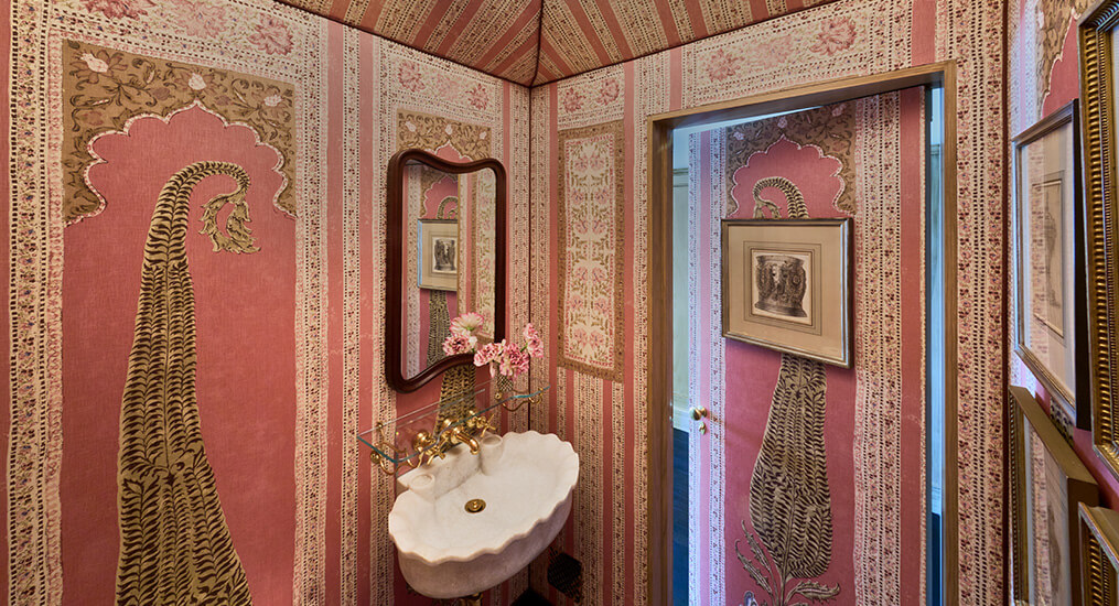 CNR Group NYC luxury home project management interior: Small bathroom with ornate pink and gold patterned wallpaper, a scalloped white sink, brass faucet, wall mirror, and framed artwork.