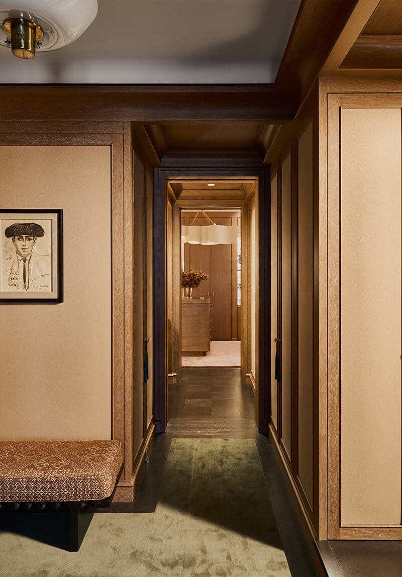 CNR Group luxury home construction interior: Narrow hallway with wood-paneled walls, dark hardwood floor, and a patterned bench on the left, leading to a room with a wooden dresser and floral arrangement.