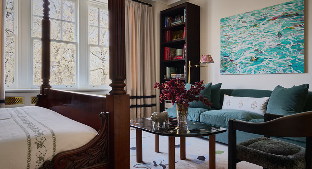 CNR Group luxury home construction interior: Cozy room with a large wooden four-poster bed, teal couch with pillows, black marble coffee table with vase of purple flowers, bookshelf, and a colorful abstract painting on the wall.