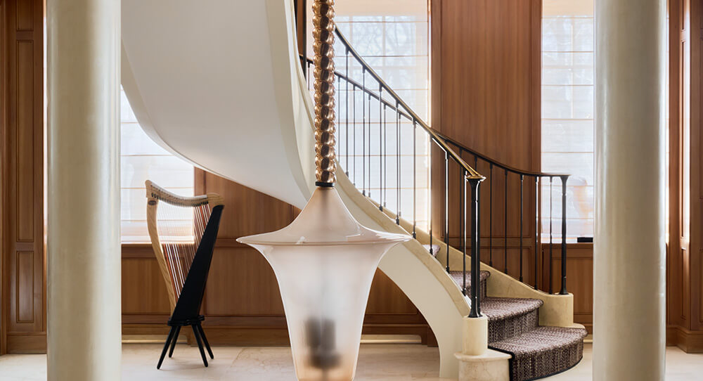 Curved carpeted staircase with black metalwork railing beside a wooden harp and white columns in a room with light filtering through window blinds.