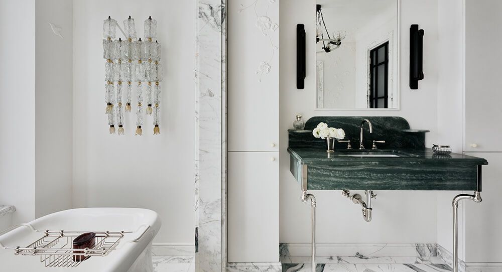 CNR Group luxury home construction interior: Bathroom with a white freestanding tub, green marble sink with chrome legs, wall mirror, and crystal wall sconces.