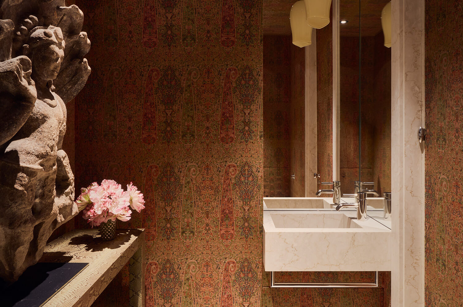 CNR Group luxury home construction interior: Bathroom with ornate patterned wallpaper, marble sink with chrome faucet, a large mirror, a wooden console table holding a vase of pink flowers, and a stone sculpture on the left.