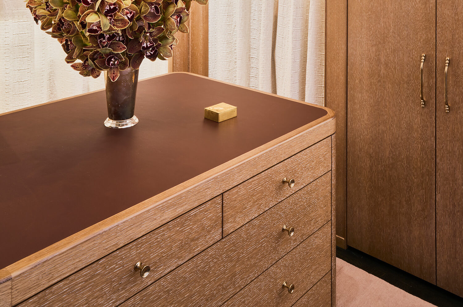 Custom millwork wooden dresser with a dark brown top, a vase of dark orchids, and a small gold box on it, next to a custom wooden cabinet with brass handles.