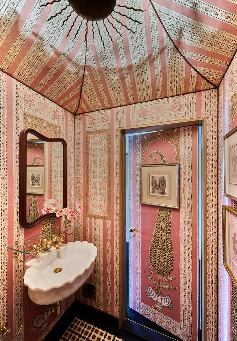 CNR Group luxury home projects management interior: Small bathroom with pink and beige patterned wallpaper and ceiling, ornate white scalloped sink with gold faucet, and framed artwork on walls and door.