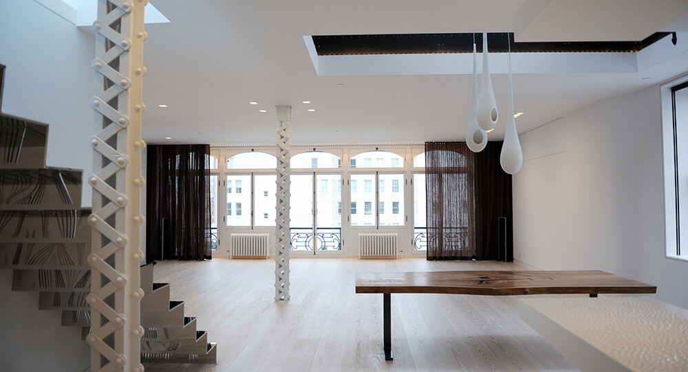 Modern, spacious room with light wood floors, large windows with black curtains, metalwork staircase, and a wooden table with hanging white pendant lights.