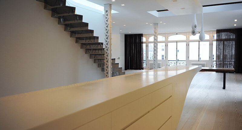 Modern interior with long white kitchen island, floating metalwork staircase, large windows with black curtains, and light wood flooring.