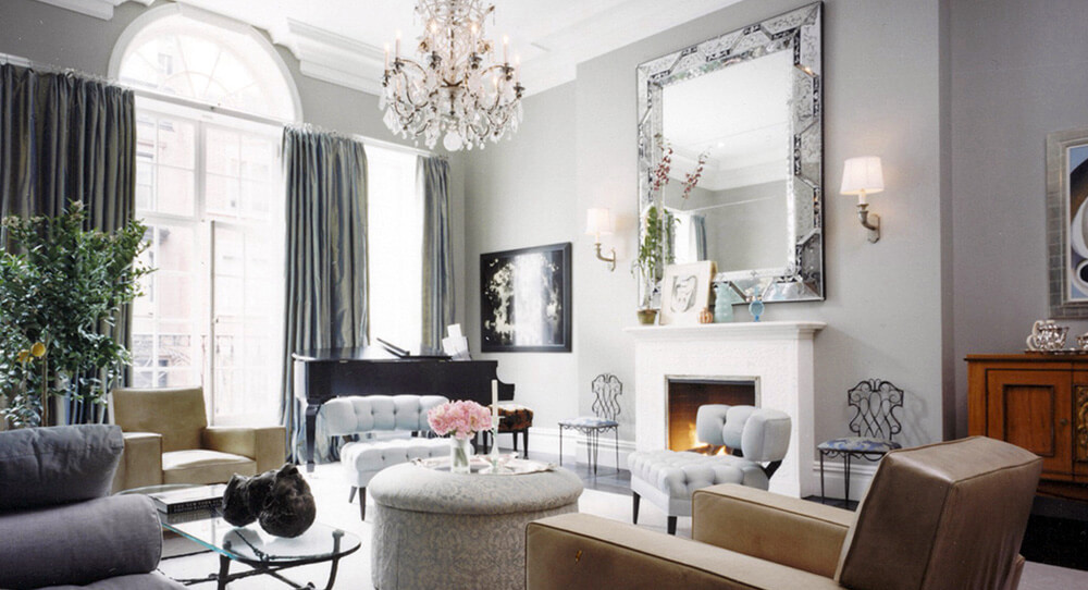 CNR Group luxury home construction interior: Elegant living room with tufted chairs, a round ottoman, a fireplace with a large mirror, and a grand piano near tall windows with dark curtains.