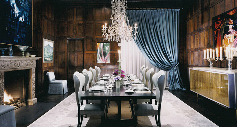 CNR Group luxury home construction interior: Elegant dining room with a long black table set for a formal dinner, white upholstered chairs, crystal chandelier, lit fireplace, custom millwork wooden paneled walls, blue drapes, and candelabras.