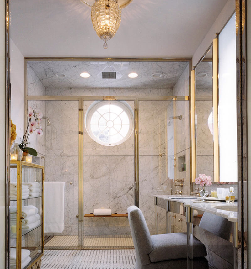 CNR Group NYC luxury home project management: Elegant bathroom with marble walls, gold-framed glass shower, a round window, and a gray upholstered chair by a vanity with dual sinks and a tall mirror.