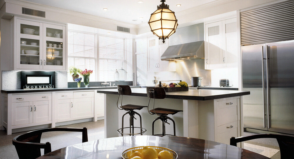 CNR Group NYC luxury home builders interior: Bright modern kitchen with white cabinets, a black countertop island, two bar stools, stainless steel appliances, and a hanging pendant light.