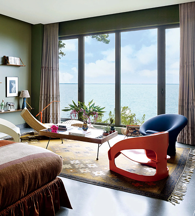 CNR Group luxury home project management interior: Modern living room with large windows overlooking a calm sea, featuring a brown bed, unique chairs, a coffee table with flowers, and neutral decor.