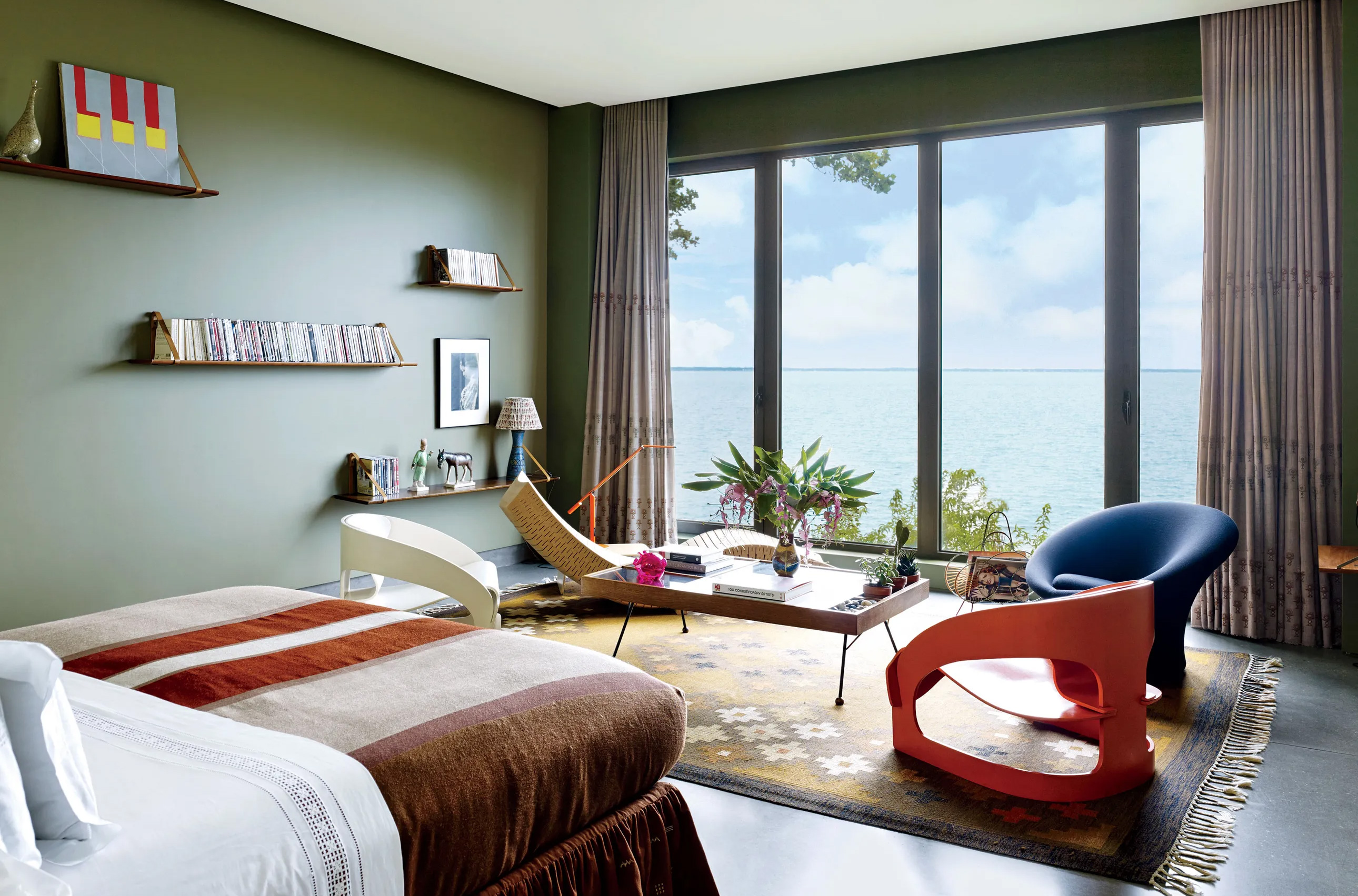 CNR Group luxury home construction interior: Cozy modern bedroom with large windows overlooking a lake, featuring a bed with brown striped blanket, unique chairs, a wooden coffee table with plants, and olive green walls with floating shelves.