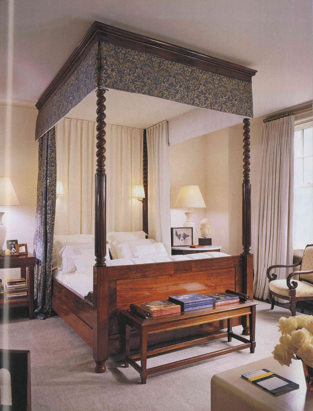 CNR Group luxury home builders project interior: Elegant bedroom with a dark wood four-poster canopy bed featuring carved posts and patterned fabric canopy, flanked by bedside tables with lamps and decorated with books, a chair, and a bouquet of white roses.