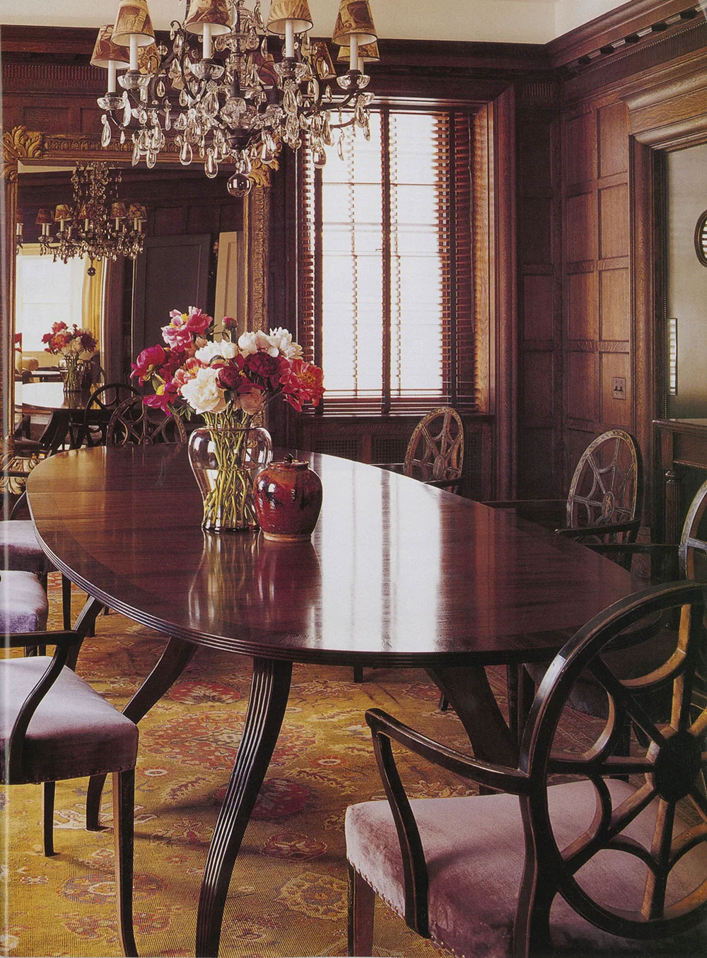 A CNR Group luxury home featuring an elegant dining room with a polished wooden table, upholstered chairs, floral centerpiece, chandelier, and wood-paneled walls with a large mirror.