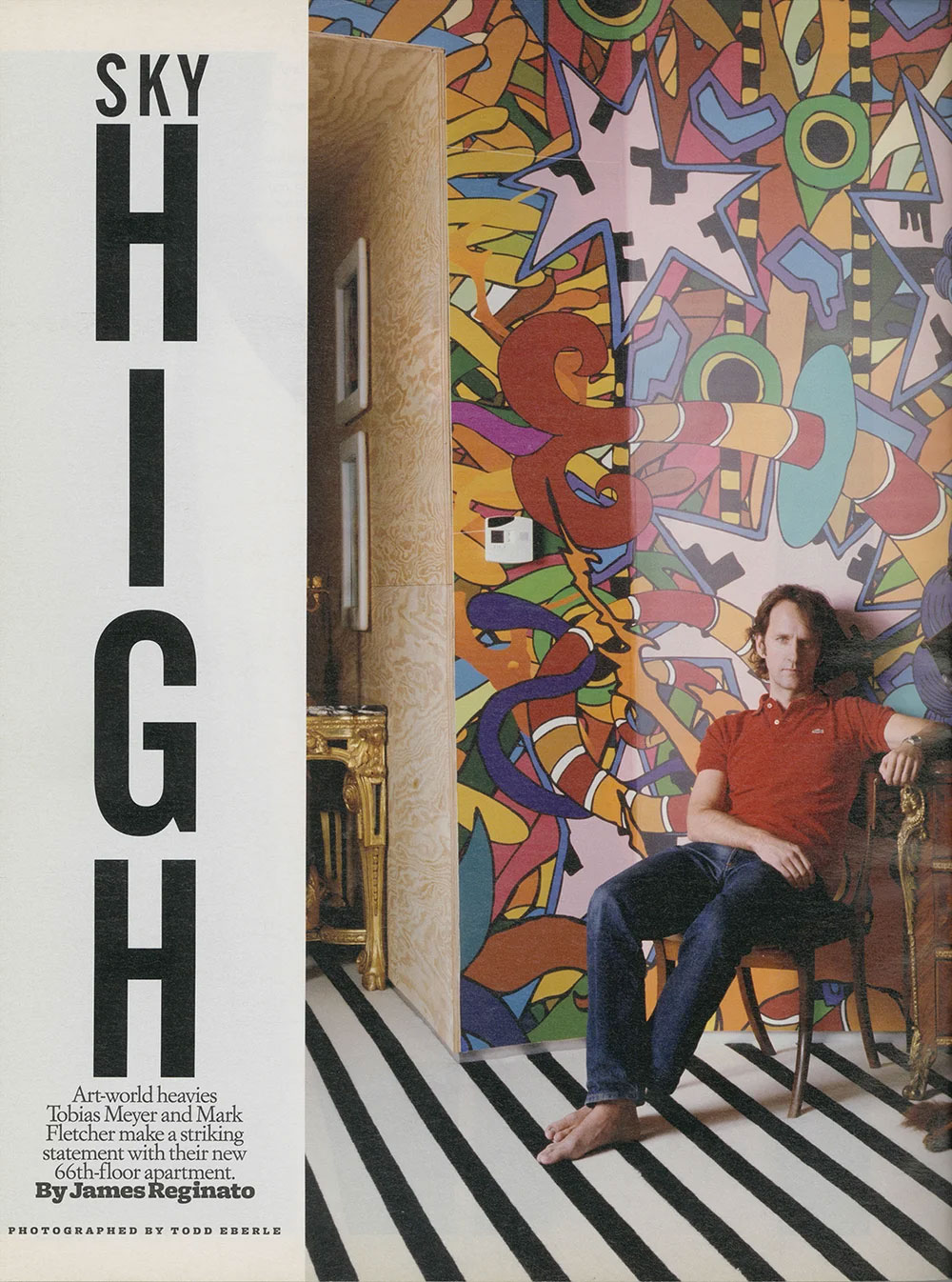 Man in a red polo shirt and jeans seated barefoot on a wooden chair against a colorful abstract mural and black-and-white striped floor in his CNR Group NYC luxury home.