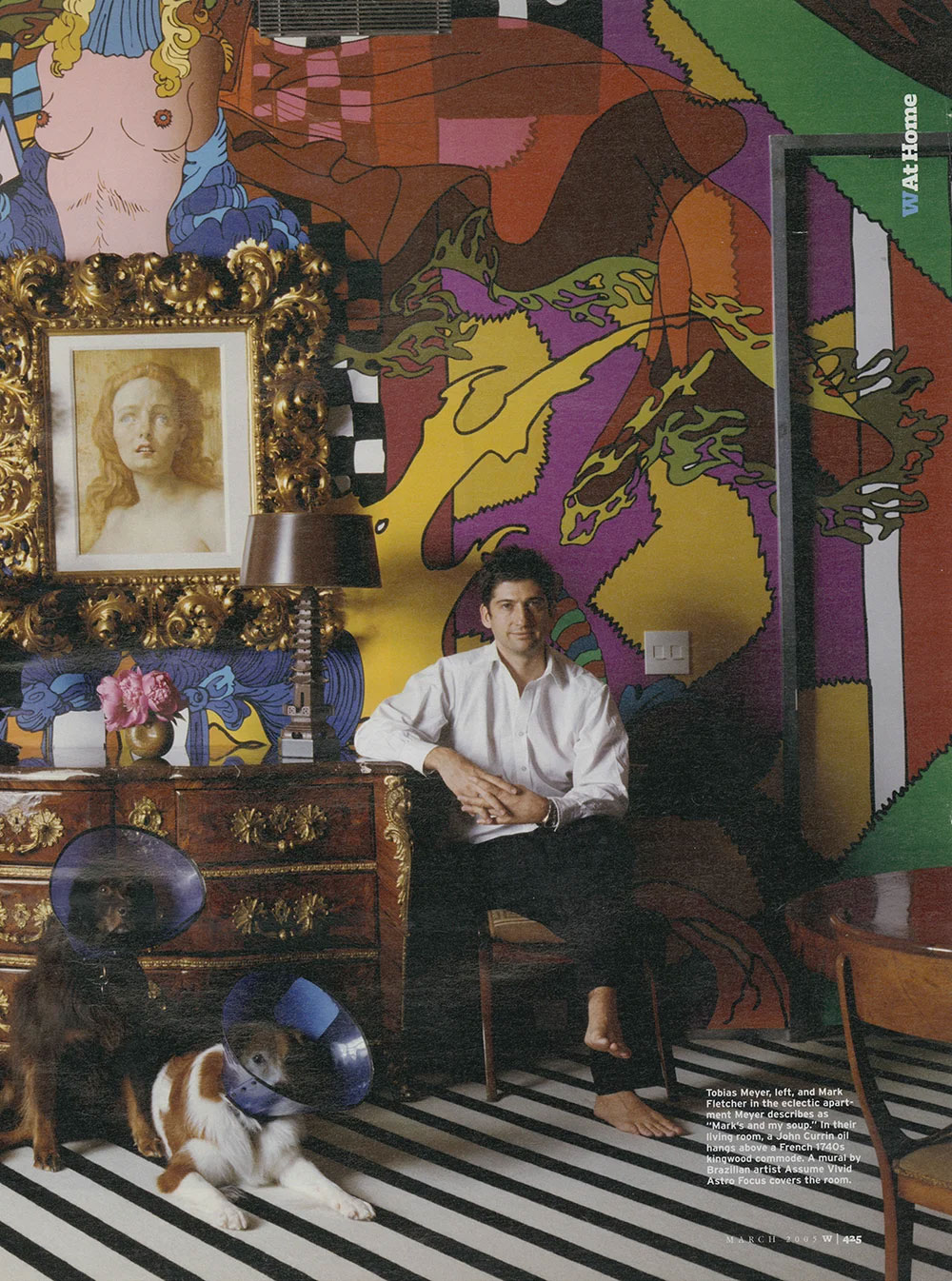 Man sitting barefoot on a chair in a colorful room of a CNR Group NYC luxury home with a striped floor, accompanied by two dogs wearing protective cones, with a large ornate framed portrait and a vibrant mural on the wall behind him.
