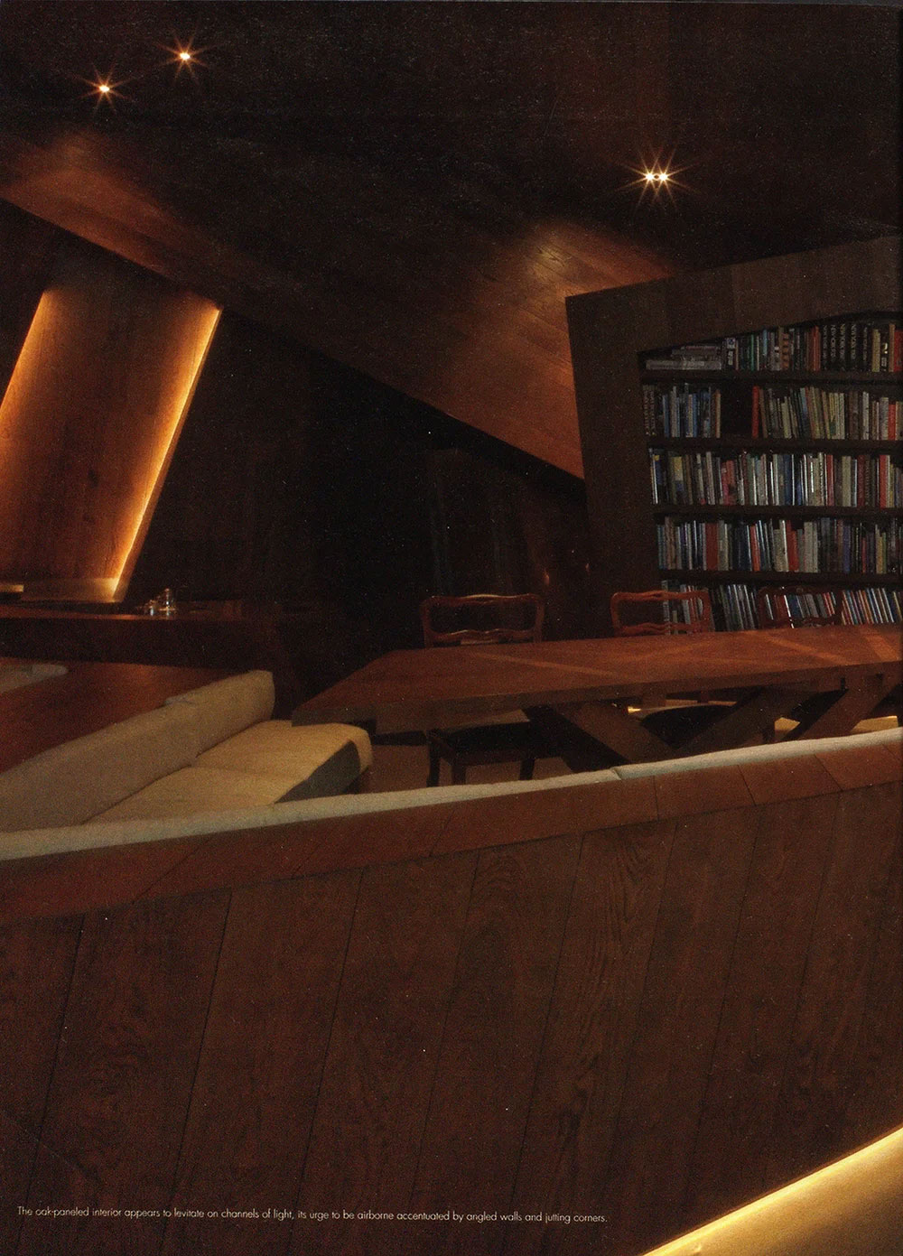 Dimly lit room within a CNR Group luxury home build with angled dark wood-paneled walls, a wooden table with chairs, a beige sofa, and a bookshelf filled with books.