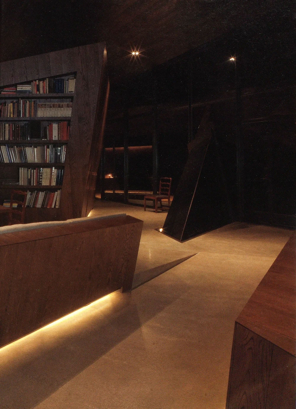 Dimly lit modern interior of a CNR Group luxury home build with wooden bookshelves, furniture, and chairs on a polished concrete floor.