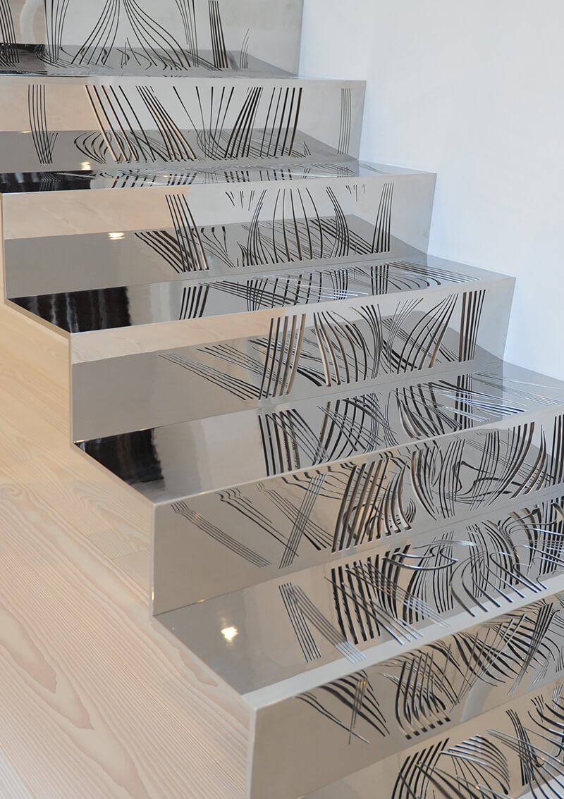 Modern staircase with custom metalwork from CNR Group that features highly reflective metallic surfaces showing black abstract line patterns reflected on each step.