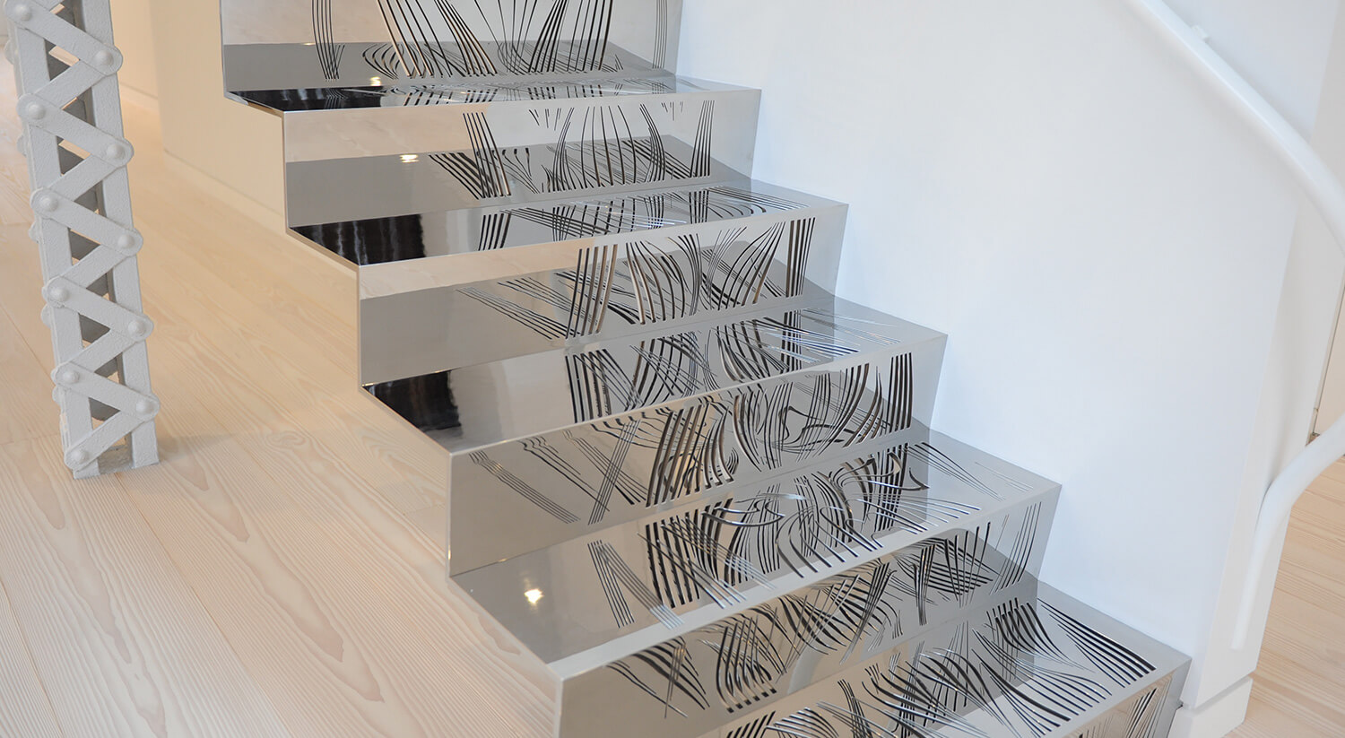 Modern staircase with custom metalwork from CNR Group featuring reflective metal steps with intricate black line patterns reflecting light in a bright room.