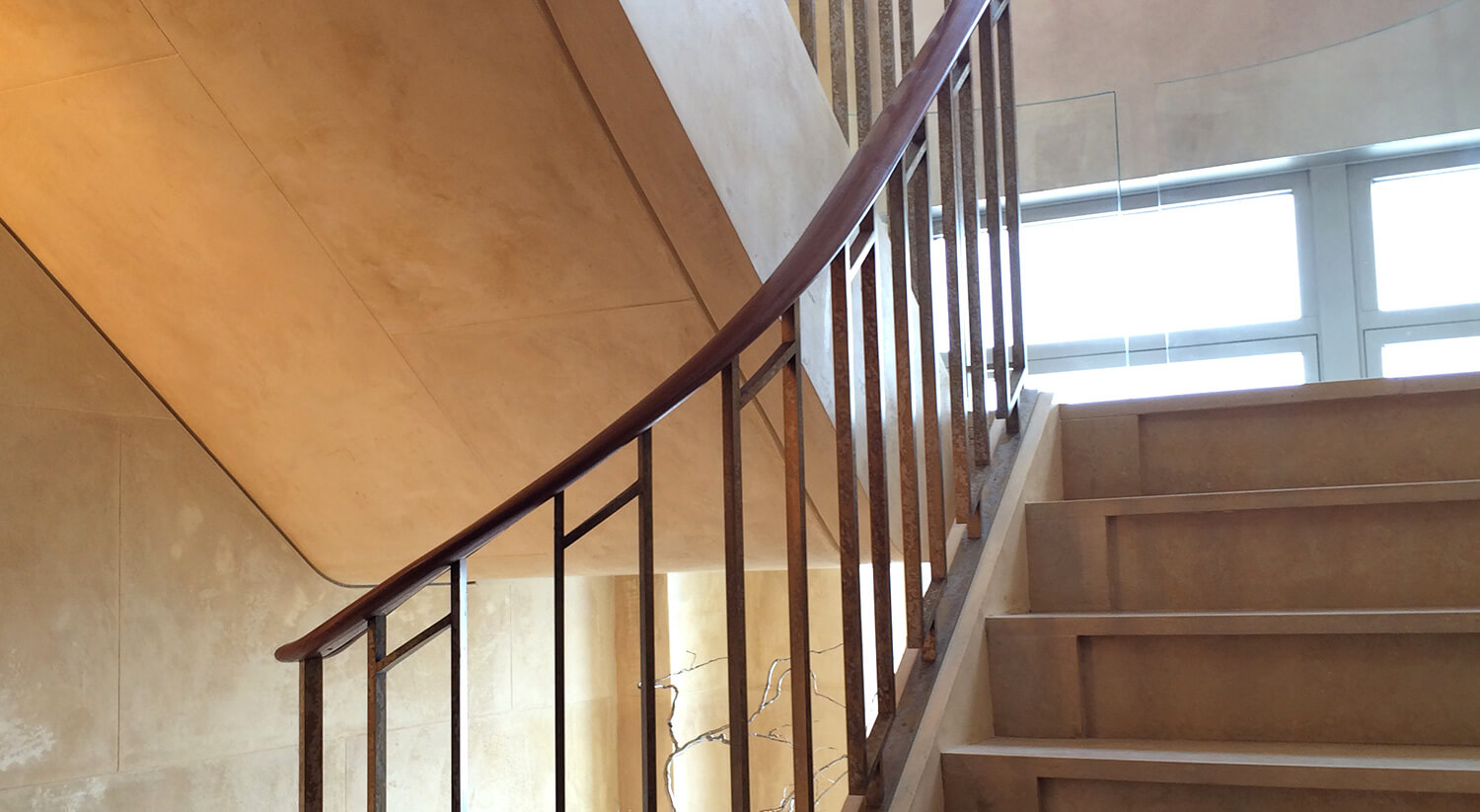 Indoor custom metalwork staircase from CNR Group with beige steps and metal railing featuring a dark wooden handrail next to a window.