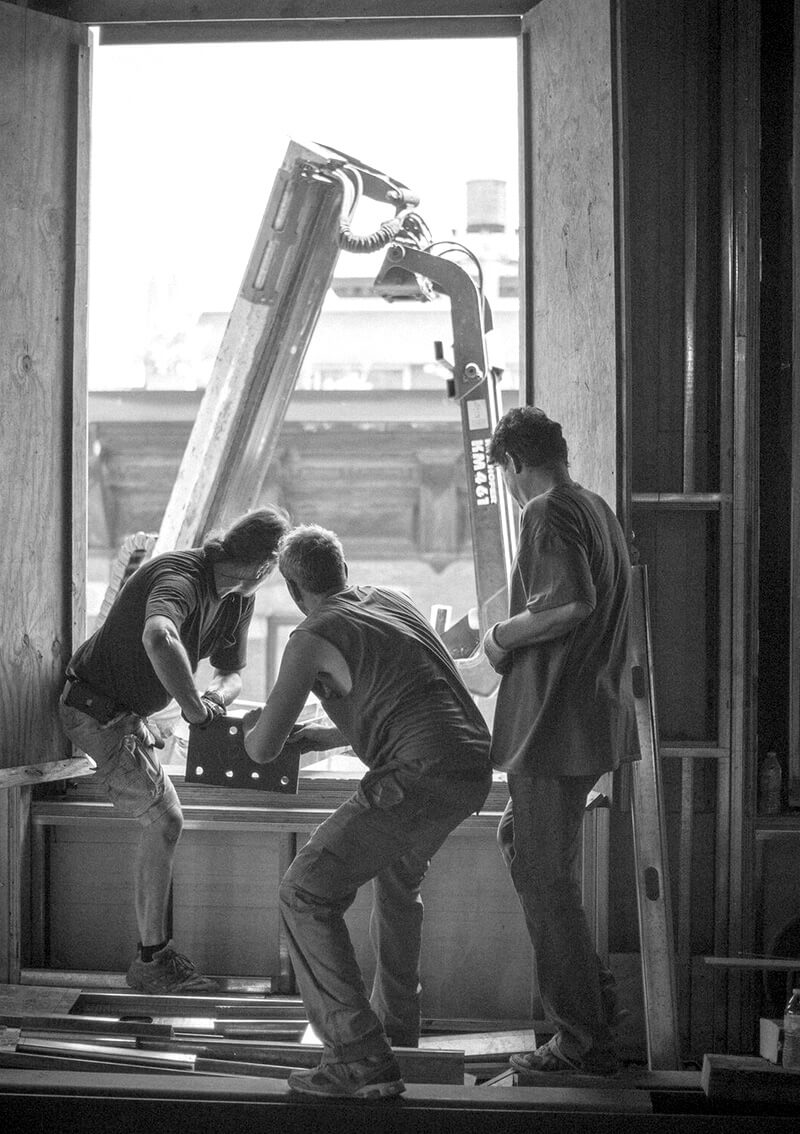 Three CNR Group NYC luxury home construction workers inside a building frame lifting or positioning a heavy metal piece near an open window with a construction crane visible outside.
