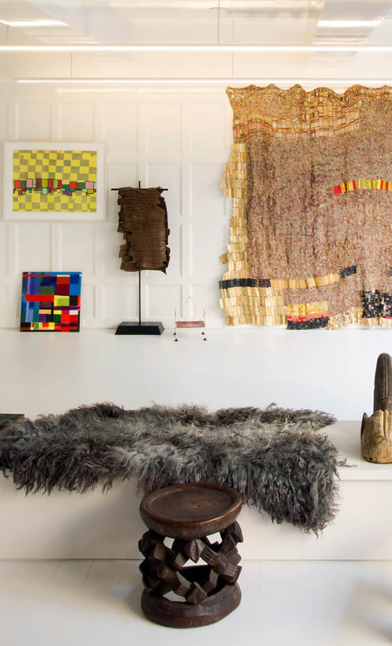 Interior of a CNR Group NYC luxury home featuring a gallery wall with colorful abstract paintings, a woven textile, wooden sculpture on white ledge, and a small carved wooden stool with a shaggy gray fur throw.