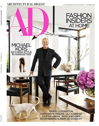 The cover of an Architectural Digest magazine that features a CNR Group luxury home build. On the cover, Michael Kors stands in a black suit standing in a modern dining room with a white table, wooden chairs, a cat walking, and large windows.