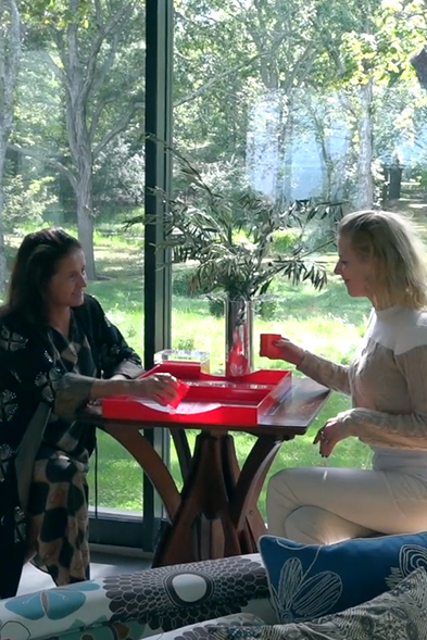 Two women sitting across from each other at a wooden table in a CNR Group luxury home build playing a game with red pieces and trays in a bright room with large windows overlooking green trees.