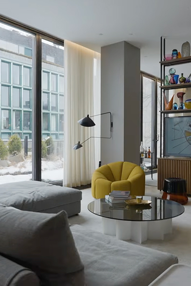 Modern living room of a luxury home in NYC with large windows, a mustard yellow curved armchair, gray sectional sofa, black wall-mounted lamp, and a round glass coffee table.