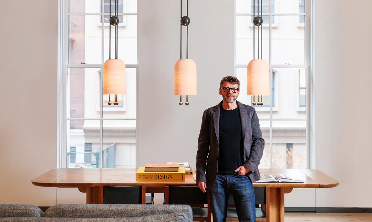 Jesse leaning on a table looking at the camera. Black t-shirt, grey jacket and jeans with a gorgeous room in the background.