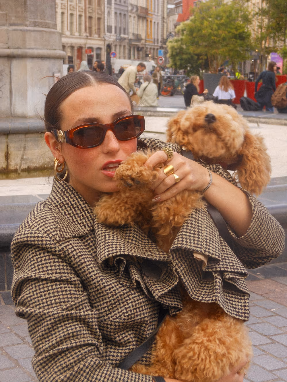 Visuel de campagne urbain montrant une femme avec des lunettes de soleil foncées et un manteau au motif pied-de-poule, tenant dans ses bras un petit chien bouclé de couleur caramel. — Campagne de lancement de la marque de vêtements pour chiens Chienne. — Design graphique par le studio Pryzma.