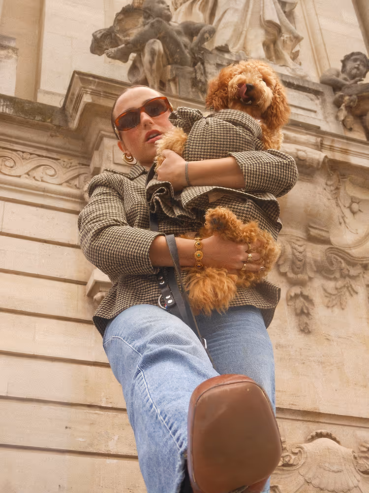 Visuel de campagne lifestyle montrant une personne stylée (veste à carreaux, lunettes de soleil) tenant un chien à poil bouclé portant un manteau assorti, posant devant une façade architecturale sculptée. — Campagne de lancement de la marque de vêtements pour chiens Chienne. — Design graphique par le studio Pryzma.