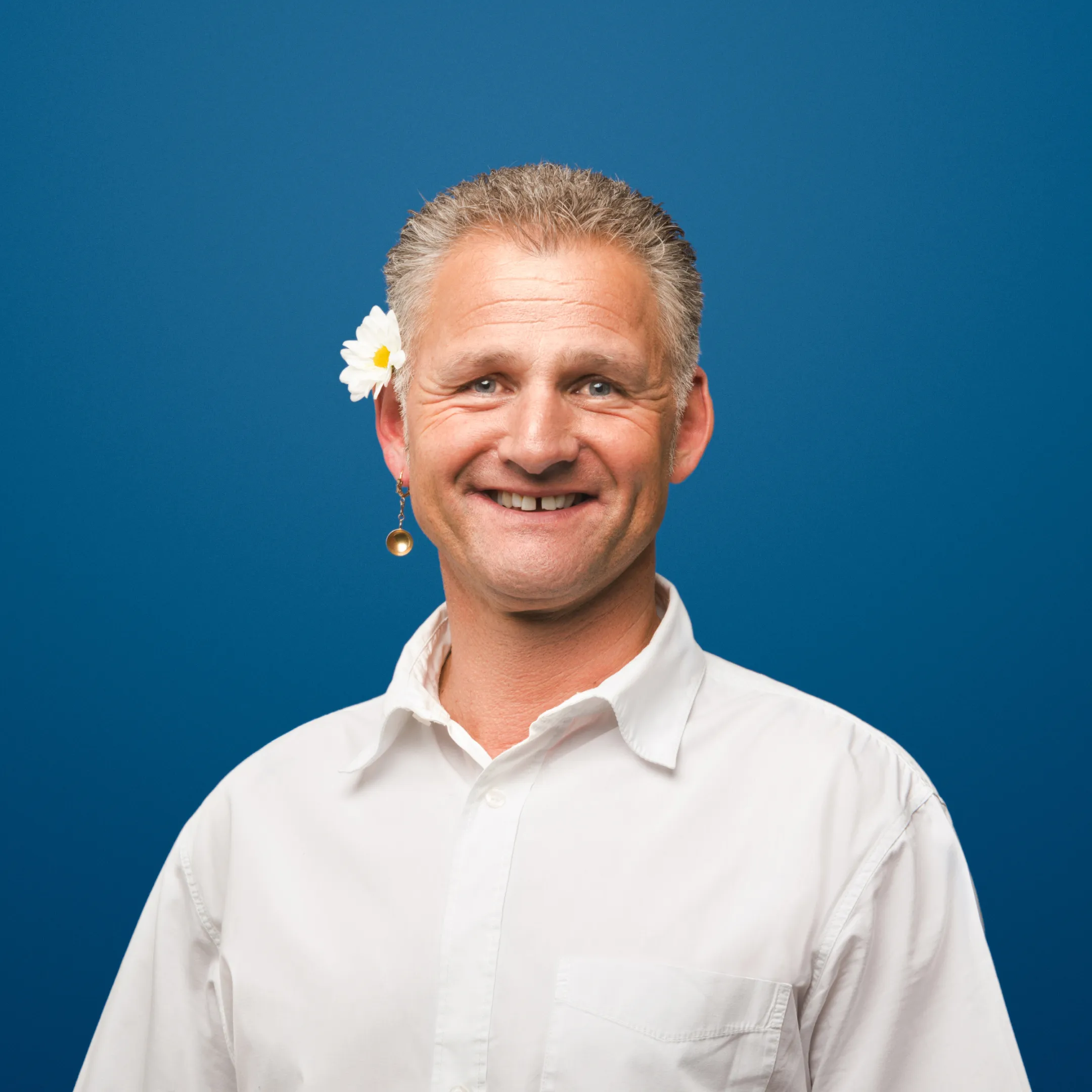 Lächelnder Mann in weissem Hemd vor blauem Hintergrund, mit Blume im Haar und Appenzeller Ohrring in Form einer goldenen Schaufel.