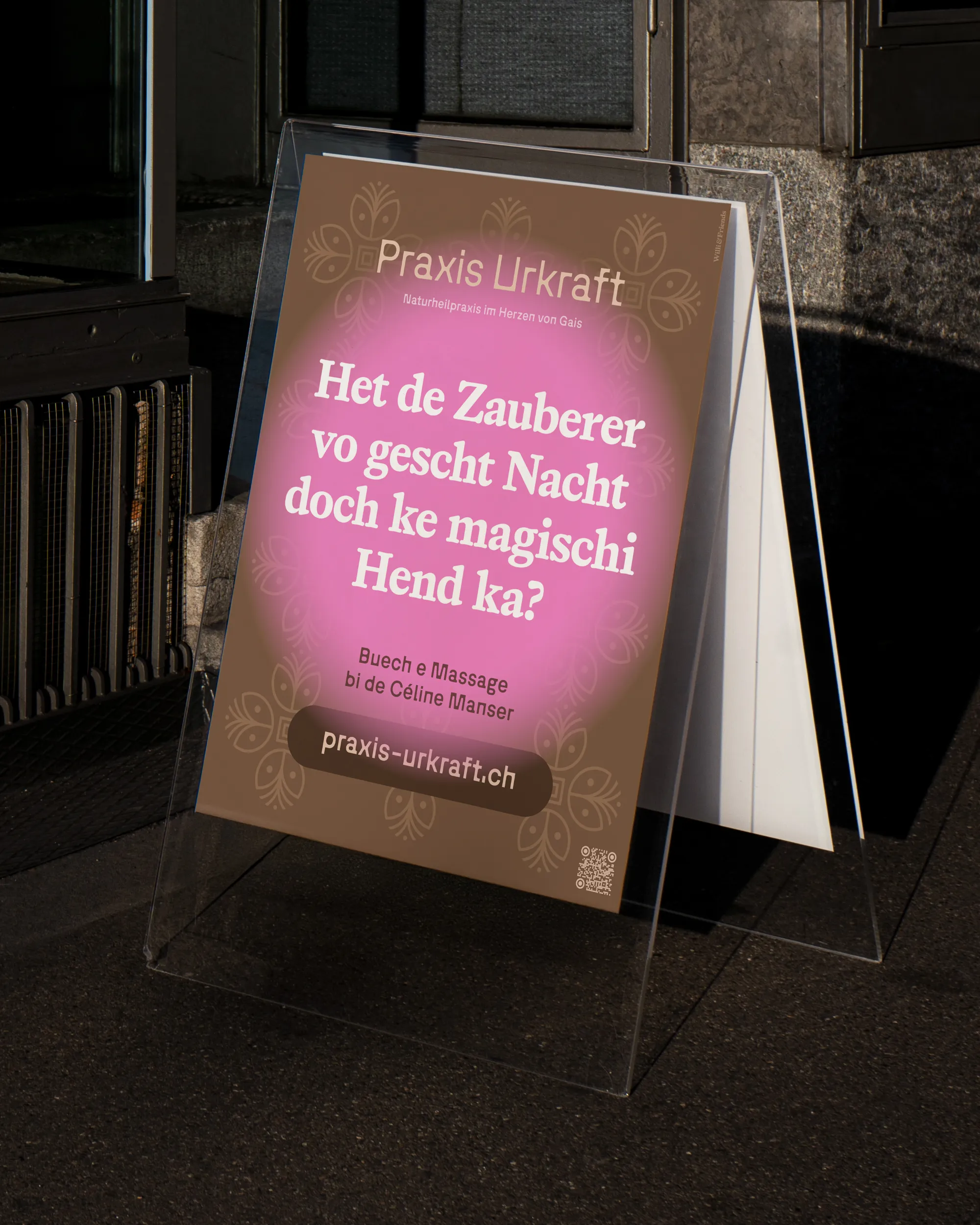 Plakatständer vor einem Gebäude mit Poster der Praxis Urkraft: brauner Hintergrund mit floralem Muster, mittig pinker Kreis mit weisser Schrift im Dialekt ‚Het de Zauberer vo gescht Nacht doch ke magischi Hend ka?‘, darunter Hinweis auf Massage bei Céline Manser und die Website praxis-urkraft.ch.