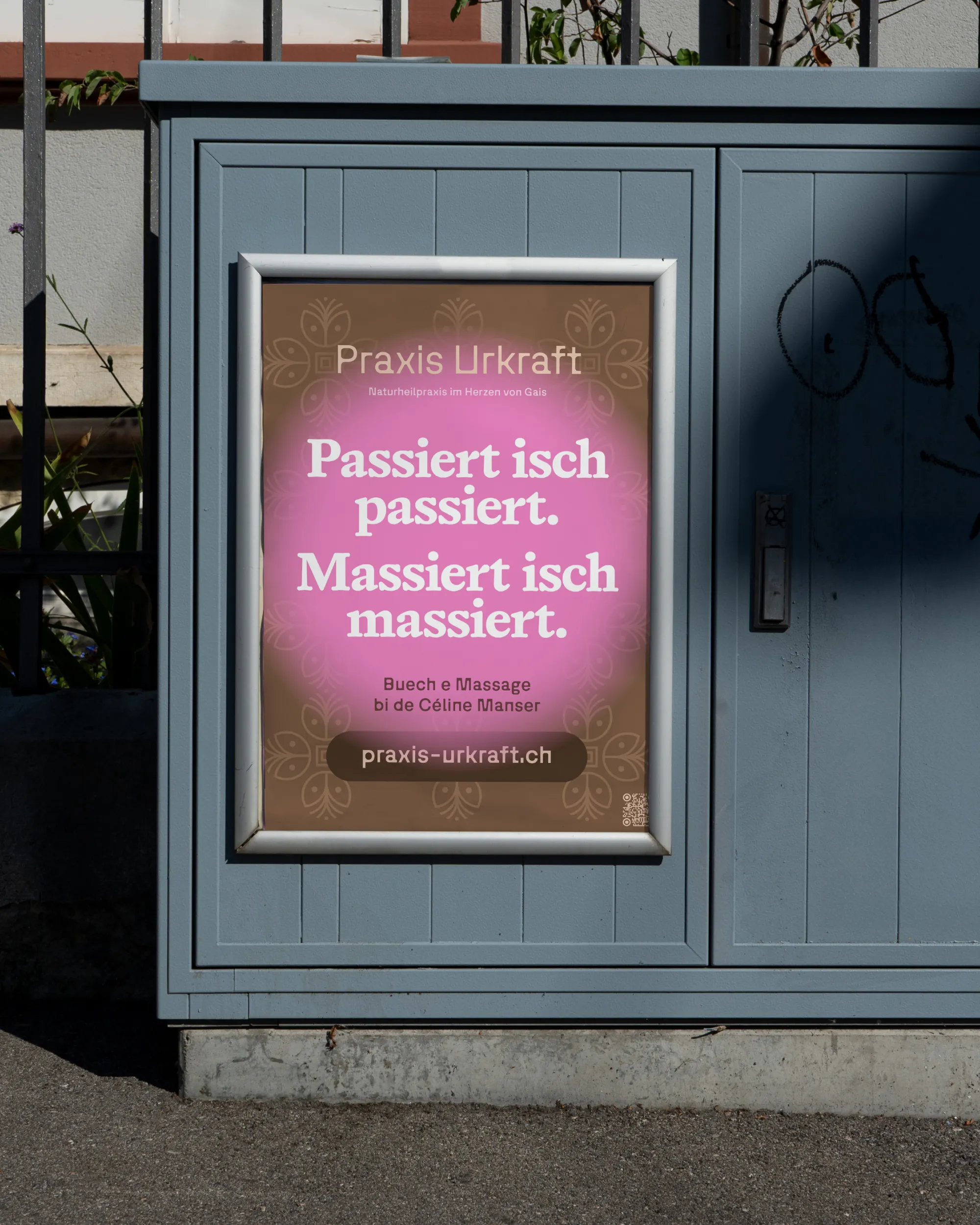 Poster der Praxis Urkraft von Céline Manser an einer grauen Wand: brauner Hintergrund mit floralem Muster, mittig pinker Kreis mit weisser Schrift ‚Passiert isch passiert. Massiert isch massiert.‘, darunter Hinweis auf Massage und Website praxis-urkraft.ch.