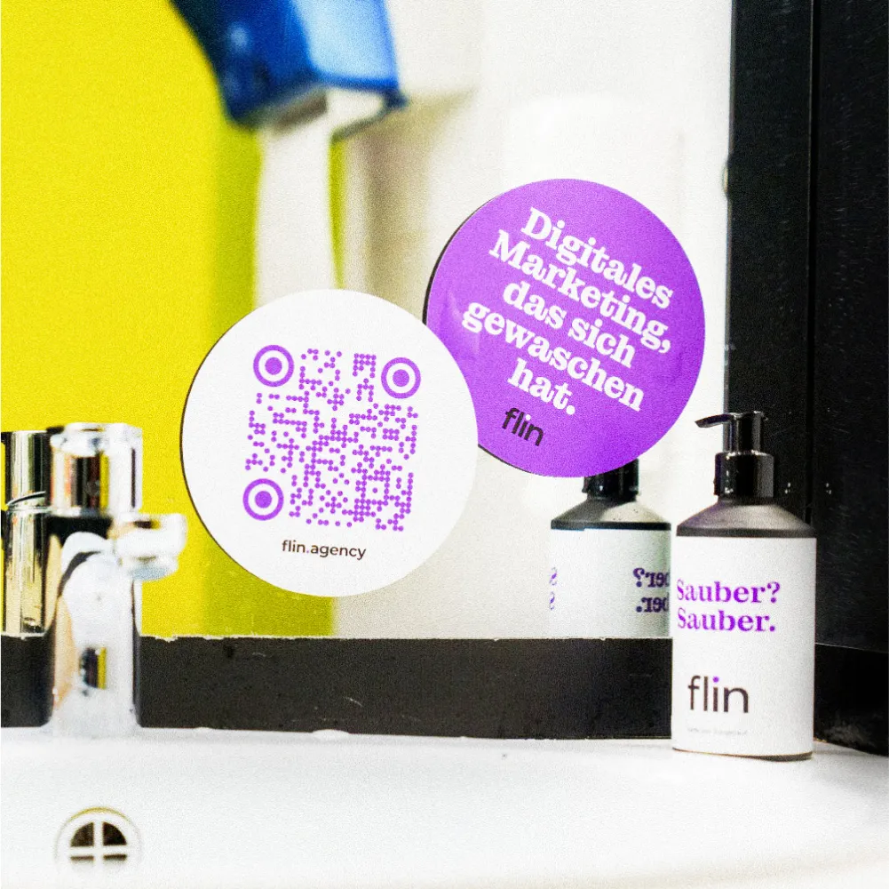 Waschbecken mit Spiegel, darauf zwei runde Sticker: violett mit der Aufschrift ‚Digitales Marketing, das sich gewaschen hat. flin‘ und weiss mit QR-Code und flin.agency; daneben ein Seifenspender mit Flin-Logo.
