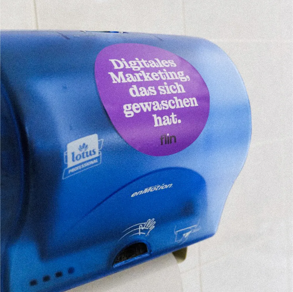 Blauer Papierhandtuchspender mit violettem Sticker der Agentur Flin, Aufschrift ‚Digitales Marketing, das sich gewaschen hat.‘
