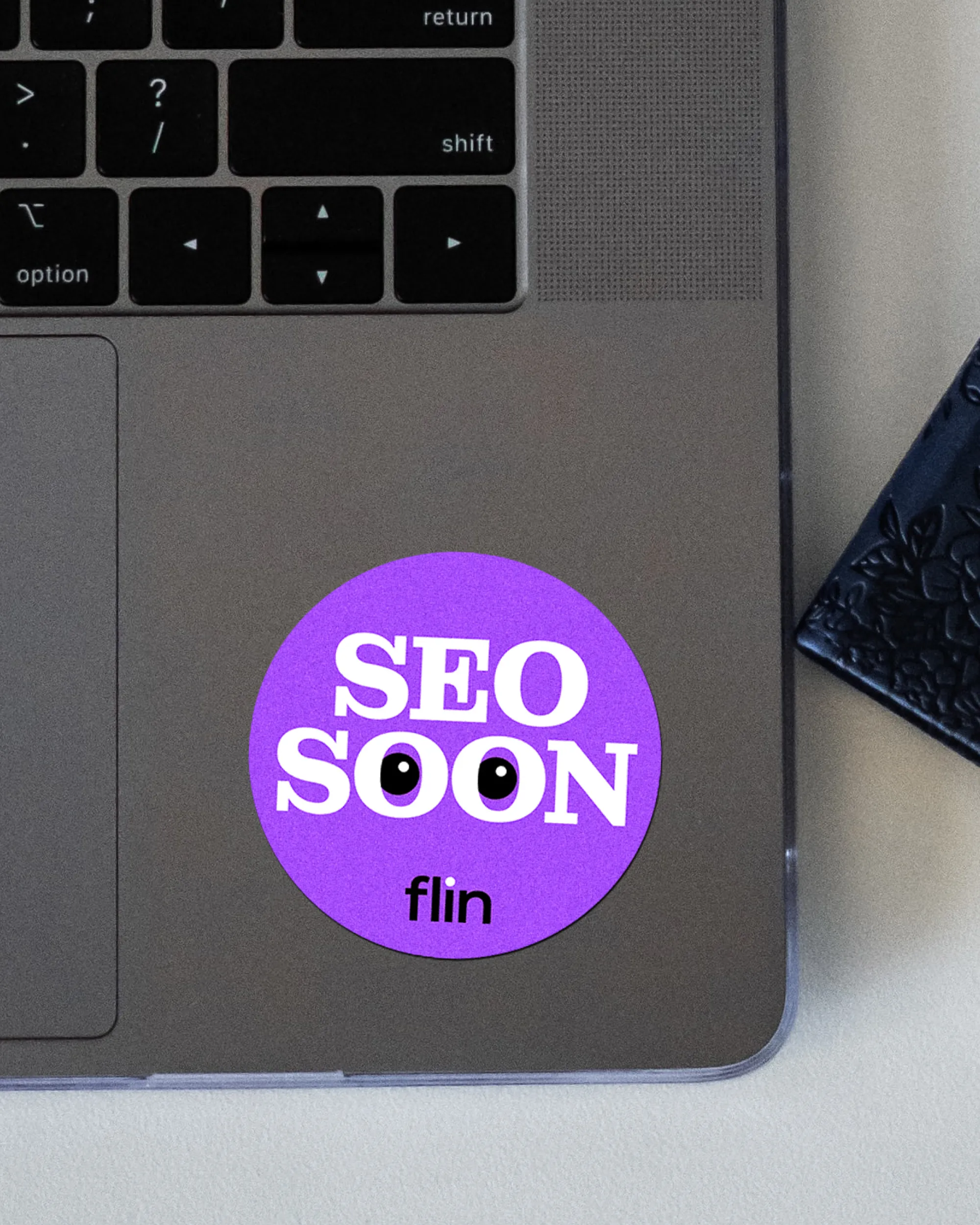 Laptop mit aufgeklebtetem runden violetten Sticker ‚SEO SOON‘ mit Augenmotiv in den Buchstaben O und Flin-Logo.