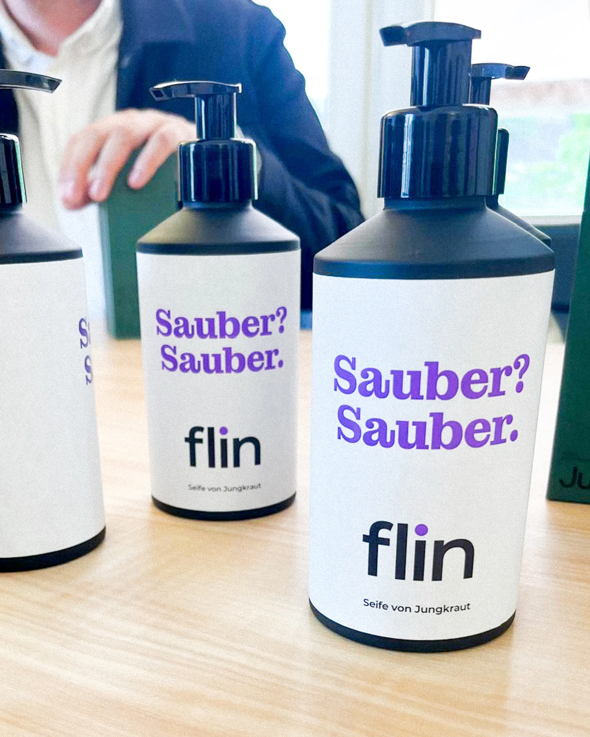 Seifenspender mit der Aufschrift ‚Sauber? Sauber.‘ und Flin-Logo auf weissem Etikett, im Hintergrund eine Person mit dunkler Jacke.