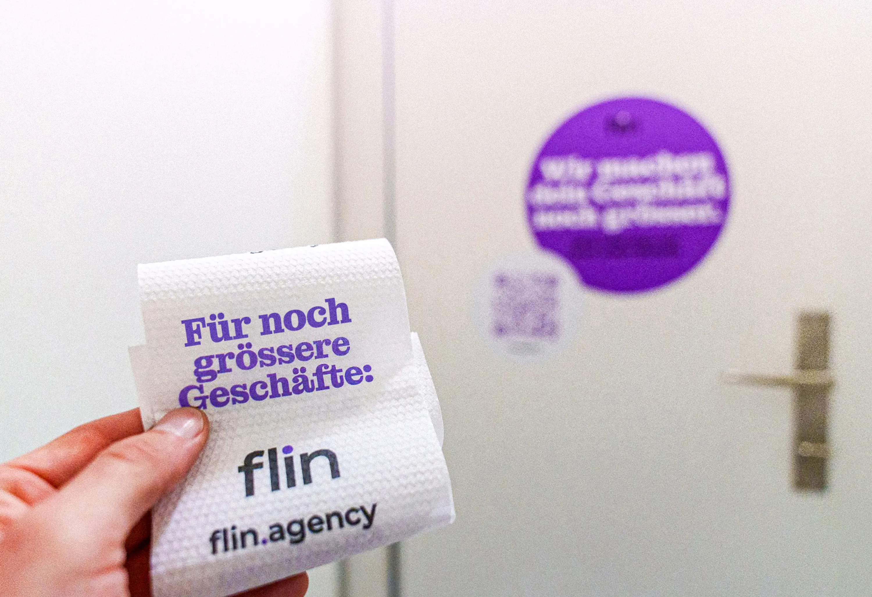 Hand hält ein Stück WC-Papier mit Aufdruck ‚Für noch grössere Geschäfte: flin.agency‘, im Hintergrund eine WC-Tür mit violettem Sticker und QR-Code.
