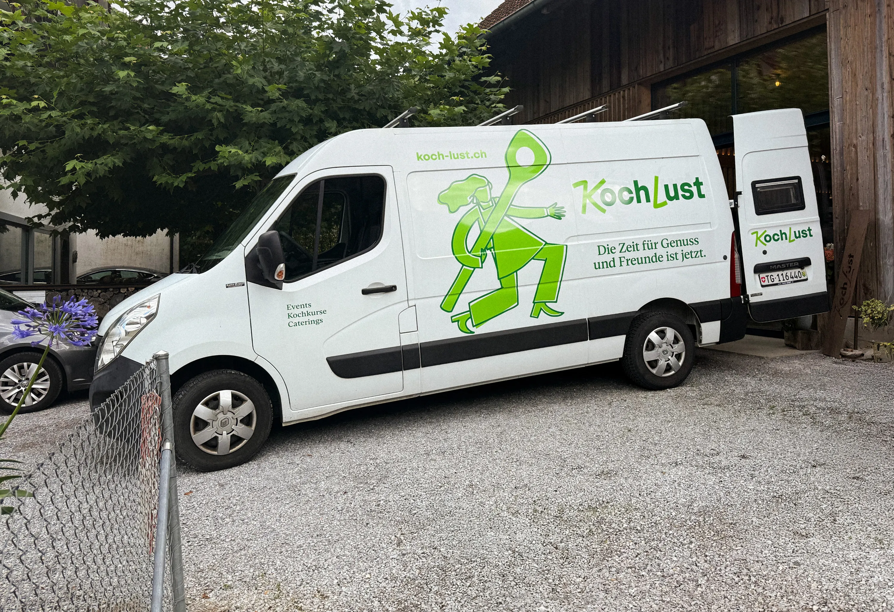 KochLust Lieferwagen mit grünem Logo und Illustrationsfigur vor Holzgebäude
